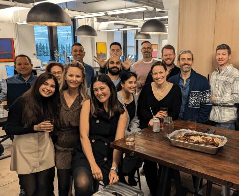 Three Media.net employees engaging in casual conversation within an office lounge area, holding coffee mugs and standing against a stylish brick wall.