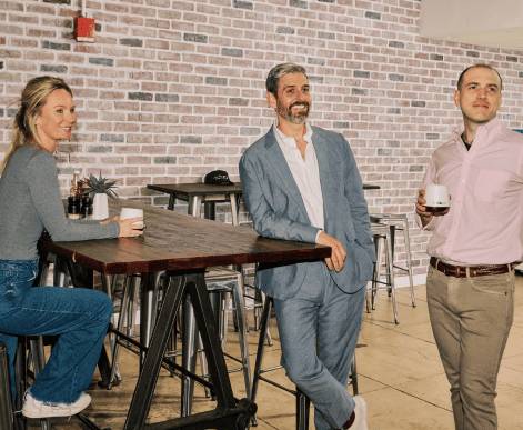 Six Media.net employees posing together outdoors at a casual company event, smiling warmly and holding beverages, against a brick wall backdrop complemented by greenery.